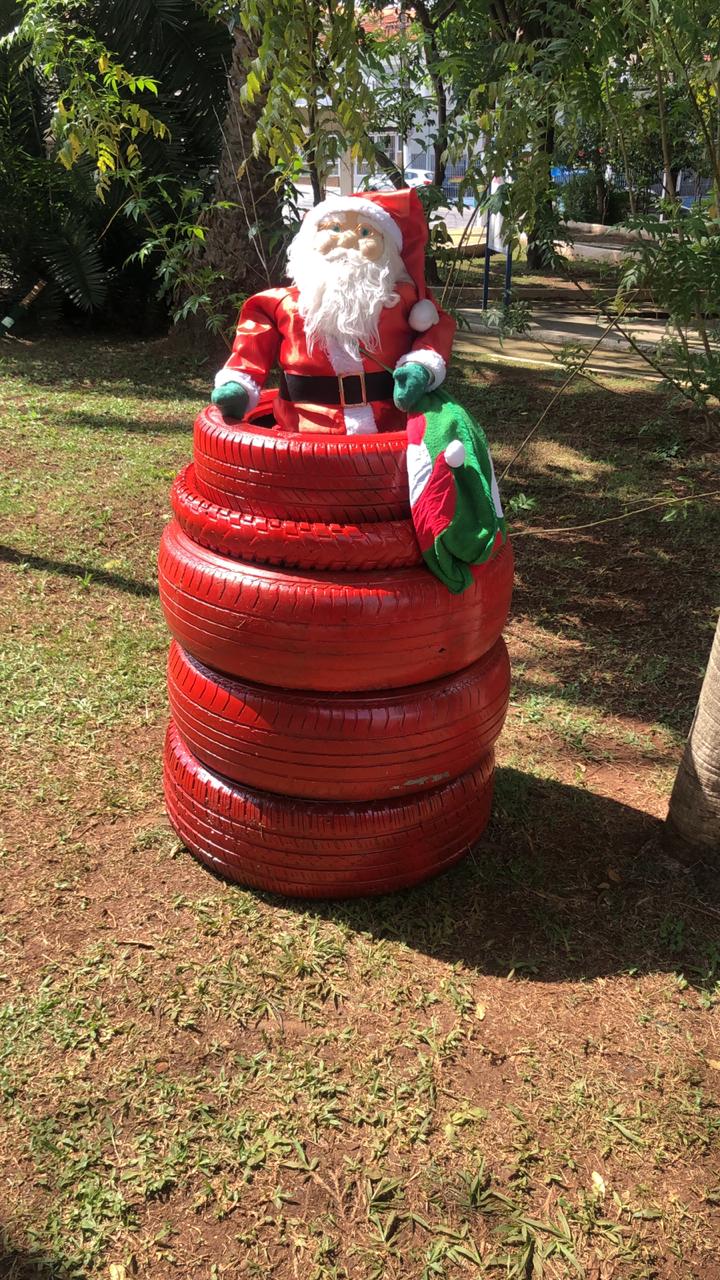 Visualiza-se o boneco de um papai noel dentro de um pneu da cor vermelha, que está em cima de outros pneus da mesma cor. A decoração está localizada em uma praça. 